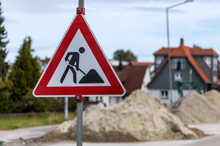 Ein Verkehrsschild, das auf eine Baustelle hinweist. Im Hintergrund befinden sich Sandhäufen auf einer Straße.